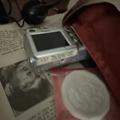 Sale Photo Thumbnail #42: Compact silver digital point-and-shoot camera shown alongside wired Sony headphones and a sealed circular patch, resting on printed newspaper.
