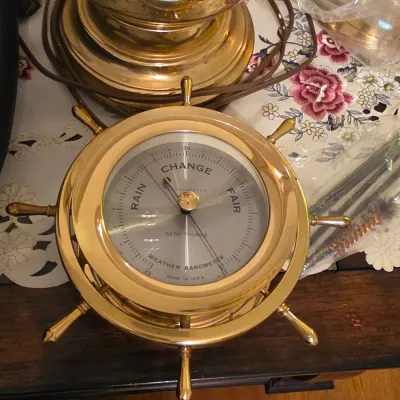 Vintage Seth Thomas brass ship's wheel weather barometer, set on a floral tablecloth alongside brass lamp components.