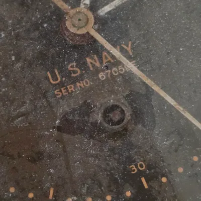 Vintage U.S. Navy military clock or gauge face with a black dial and serial number 67059, showing light wear and surface dust.