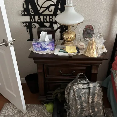 Dark wood nightstand with drawer and door, holding a vintage hurricane-style lamp, clear picture frame, brass bell, tissue box, and...