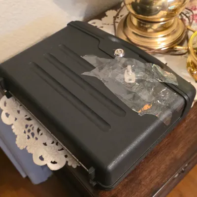 Black molded plastic lockbox with keys taped to the lid, displayed on a wooden table beside brass decor.