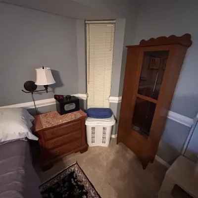 Bedroom corner featuring a tall wooden cabinet, a three-drawer wooden nightstand topped with a small digital safe, and a wall-mounted...