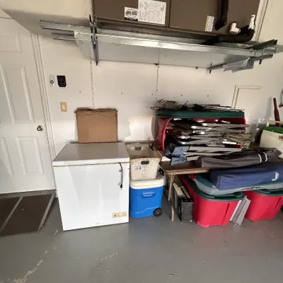 White compact chest freezer, blue rolling cooler, plastic storage totes, and folding lawn chairs arranged against a garage wall.