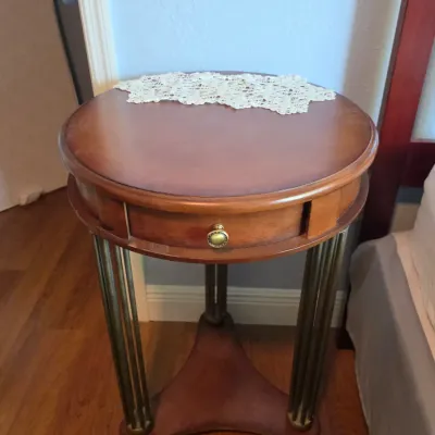 Round wood side table featuring a single drawer with brass hardware, reeded metal legs, and a triangular lower stretcher base, topped...