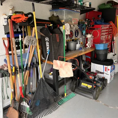 Assortment of garage items including a gas-powered lawn mower, long-handled yard tools, rakes, shovels, and a red rolling tool chest.