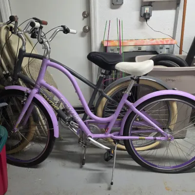 Electra Townie cruiser bicycle in purple and grey, featuring a step-through frame, white padded saddle, matching fenders, and kickstand.
