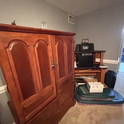Large wooden armoire with arched panel doors and bottom drawers, standing alongside a computer desk in a bedroom setting.