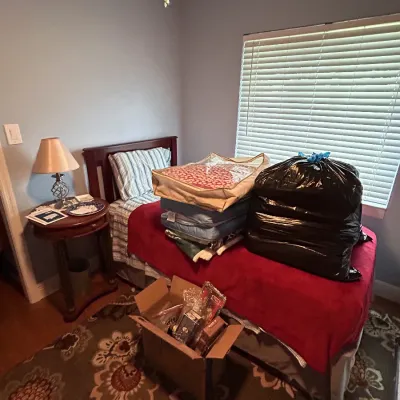 Bedroom featuring a dark wood twin bed frame, round side table with lamp, patterned area rug, and various packed linens and household items.