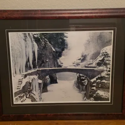 Framed winter landscape photograph depicting a stone bridge and frozen gorge, set in a dark double mat and rich burl wood-style frame.
