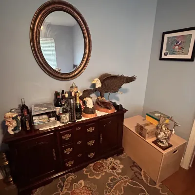 Traditional dark wood sideboard displaying liquor bottles and eagle sculptures beneath a dark oval wall mirror.