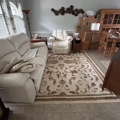 Living room setup featuring a cream-colored leather sofa and matching recliner, a large floral area rug, end tables, and moving boxes...