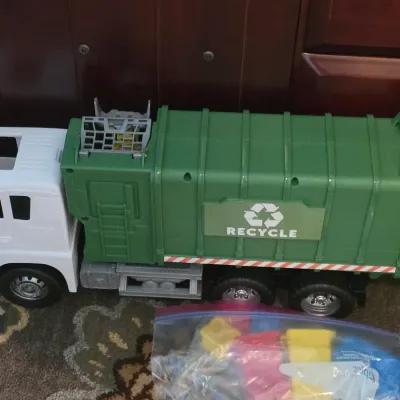 Large plastic toy recycling truck in green and white, accompanied by a plastic bag filled with colorful mini trash bins.