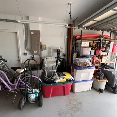 Garage storage area featuring plastic storage totes, a hand truck, a purple Townie bicycle, and various household utility items.