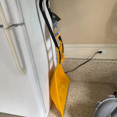 Sale Photo Thumbnail #129: Yellow SnoPlow ergonomic snow shovel with a metal edge and curved black handle, stored next to a refrigerator.