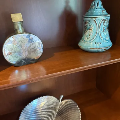 Sale Photo Thumbnail #51: Collection of decorative home accents, including a metallic art glass decanter, a distressed turquoise ceramic jar, and a silver-tone leaf-shaped dish, displayed on wooden shelves.