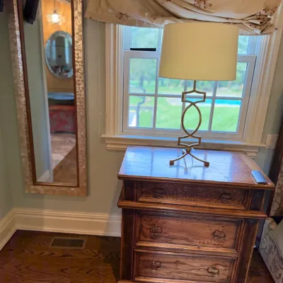 Sale Photo Thumbnail #96: Rustic three-drawer wooden nightstand topped with a contemporary geometric brass table lamp, paired with a full-length mosaic-framed wall mirror.