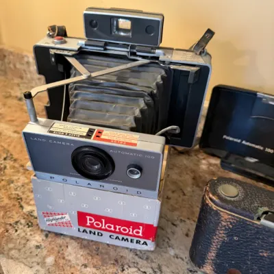 Sale Photo Thumbnail #190: Vintage Polaroid Automatic 100 Land Camera with expanding bellows, resting on a Polaroid box alongside its protective hard shell case and a worn leather camera case.