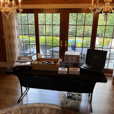 Sale Photo Thumbnail #119: Display table with various electronics including a Crosley turntable, boxed Apple TV, drone equipment, and a plastic storage bin filled with cables.
