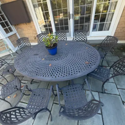 Sale Photo Thumbnail #21: Large dark grey cast aluminum outdoor dining set featuring a large round lattice-top table and matching armchairs, set on a stone patio.