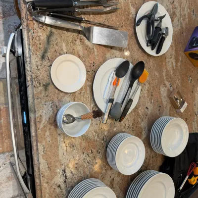 Sale Photo Thumbnail #135: Assorted white ceramic dinnerware stacks paired with barbecue utensils, kitchen gadgets, and serving spoons on a granite countertop.