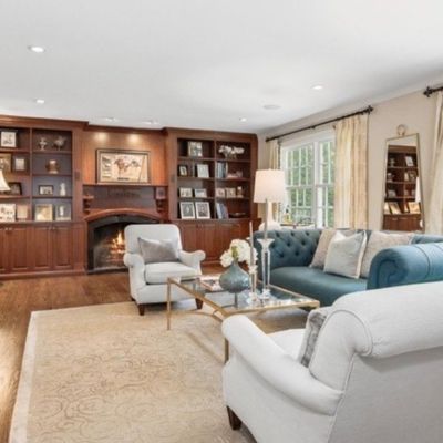 Sale Photo Thumbnail #4: Spacious living room featuring a teal tufted Chesterfield sofa, white upholstered armchairs, glass-top coffee table, and custom built-in wooden shelving around a fireplace.