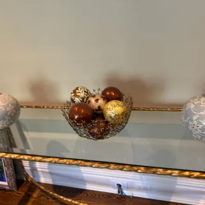 Sale Photo Thumbnail #53: Gilt metal twisted-rope console table with a glass top, flanked by a pair of pale blue floral ceramic vases and a wire centerpiece bowl filled with decorative orb accents.