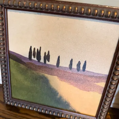 Sale Photo Thumbnail #117: Framed minimalist landscape print depicting cypress trees on rolling hills, housed in a beaded brown and gold-toned frame.