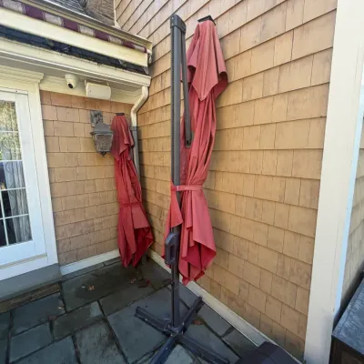 Sale Photo Thumbnail #22: Pair of large cantilever patio umbrellas with dark metal frames and red fabric, standing folded on a stone patio.
