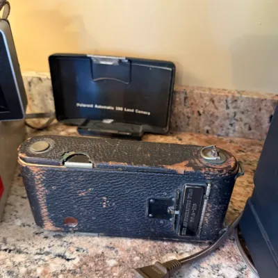 Sale Photo Thumbnail #192: Vintage Kodak No. 1A Autographic Kodak Jr. folding camera with worn black leatherette casing, displayed alongside a Polaroid Automatic 100 Land Camera cover.