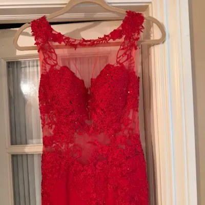 Sale Photo Thumbnail #109: Red formal evening gown featuring an illusion lace bodice with sequin detailing, hanging on a beige hanger against a door frame.