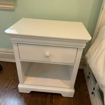 Sale Photo Thumbnail #168: White wooden nightstand featuring a single upper drawer with a round knob and an open lower storage shelf.