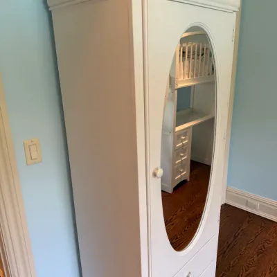 Sale Photo Thumbnail #155: White painted wooden armoire featuring an oval mirror on the front door, a molded top crown, and lower storage drawers.
