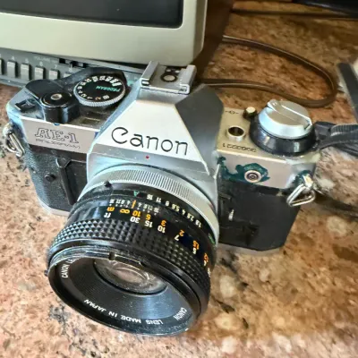 Sale Photo Thumbnail #188: Vintage Canon AE-1 Program 35mm SLR camera fitted with a Canon lens, showing visible surface wear and tear.
