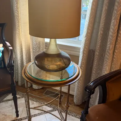 Sale Photo Thumbnail #60: Modern glass table lamp with a beige drum shade, resting on a round glass-top metal accent table flanked by wood-trimmed armchairs.