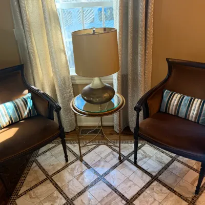 Sale Photo Thumbnail #56: Pair of vintage French-style armchair seats with dark brown upholstery, paired with a brass round glass-top side table and a ceramic table lamp.