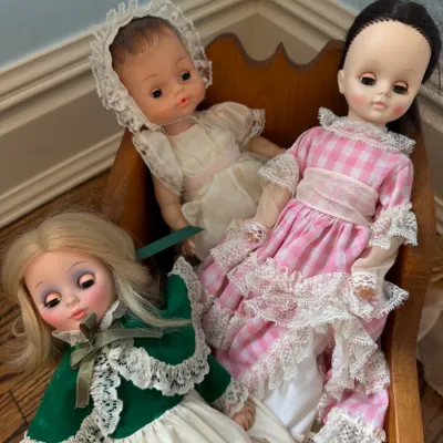 Sale Photo Thumbnail #152: Trio of vintage vinyl dolls with sleep eyes, dressed in lace-trimmed outfits and seated in a small wooden chair.