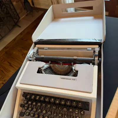 Sale Photo Thumbnail #123: Vintage Underwood 310 manual typewriter housed in its original cream and black carrying case, featuring dark keys and a compact design.