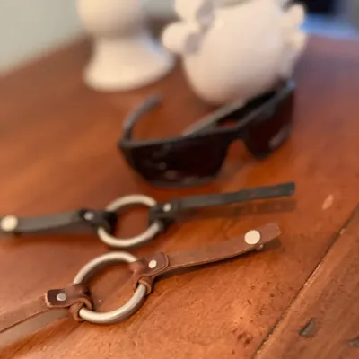 Sale Photo Thumbnail #95: Two leather O-ring wrist cuffs in brown and black leather, shown alongside a pair of dark sunglasses on a wooden surface.