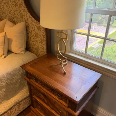 Sale Photo Thumbnail #82: Contemporary gilded openwork table lamp with a cream drum shade, seated atop a traditional wooden nightstand with brass hardware.