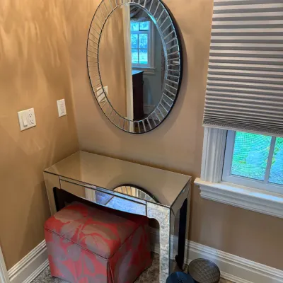 Sale Photo Thumbnail #112: Contemporary mirrored vanity desk paired with an oval wall mirror featuring a segmented frame, accompanied by an upholstered red floral ottoman underneath.