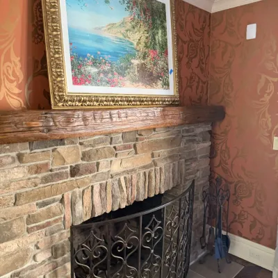 Sale Photo Thumbnail #68: Large framed Mediterranean landscape art print resting on a rustic wooden mantelpiece above a stone fireplace, complete with a scrollwork iron fireplace screen.