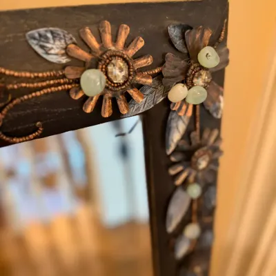 Sale Photo Thumbnail #144: Close-up of a decorative dark wooden mirror frame adorned with copper metal flower appliques, leaves, and pale green stone accents.