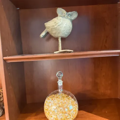Sale Photo Thumbnail #46: Stylized metallic bird figurine and a round glass decanter filled with decorative gold and amber foil chips, displayed inside a wooden bookcase.