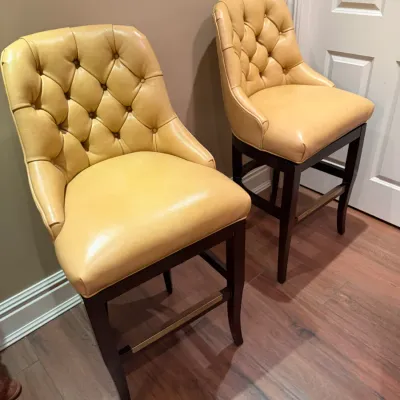 Sale Photo Thumbnail #139: Pair of modern tufted leather barstools in a buttery yellow upholstery with dark wood legs and brass footrests.