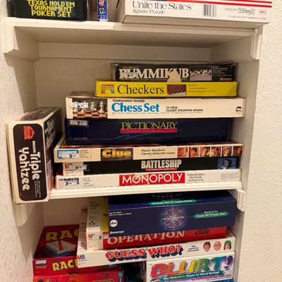 Sale Photo Thumbnail #62: Collection of classic board games, jigsaw puzzles, and card sets stacked on white built-in shelves, including Monopoly, Clue, Battleship, and Yahtzee.