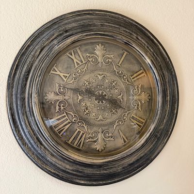 Sale Photo Thumbnail #18: Large ornate round wall clock featuring a distressed dark frame, a metallic face with embossed traditional scrolls, and bronze Roman numerals.