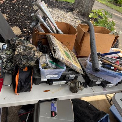 Sale Photo Thumbnail #87: Assorted garage and workshop items including hand tools, air horns, camo fabric, Evapo-Rust, and plastic storage containers arranged on a folding table.