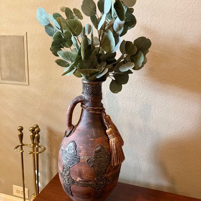 Sale Photo Thumbnail #88: Rustic terracotta jug-style vase featuring embossed metal appliqués, a single handle, and gold tassel trim, filled with artificial eucalyptus branches.