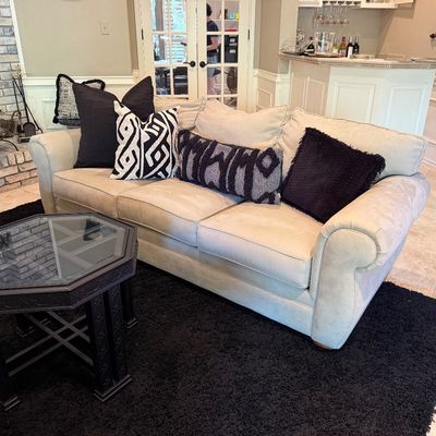 Sale Photo Thumbnail #36: Contemporary beige three-cushion sofa styled with patterned throw pillows, paired with an octagonal dark wood glass-top side table on a black shag rug.