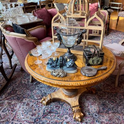 Sale Photo Thumbnail #38: Round burl wood pedestal table featuring carved claw feet, displaying an assortment of vintage items including etched glassware, a large wooden birdcage, and decorative tabletop accents.